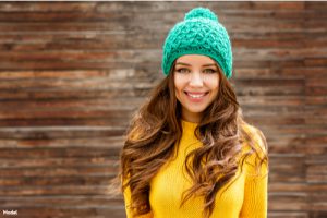 Woman with brown wavy hair in a yellow knit sweater and blue knit hat