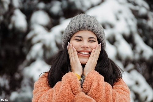 Joyful woman in a knit hat and fluffy coat gently touching her cheeks outdoors in winter