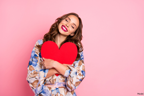 Woman with long brown hair in a flowy blue and brown blouse holding a big red paper heart to her chest and smiling