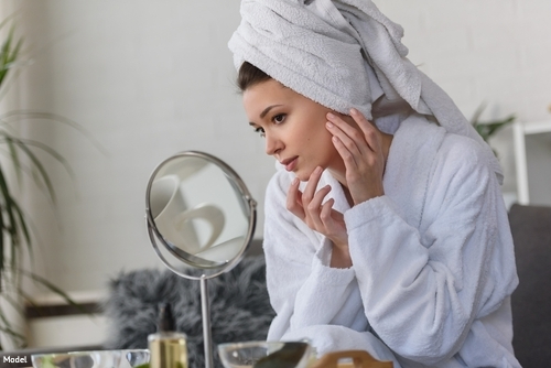 Woman wearing a robe with her hair in a towel looking at her skin in a mirror